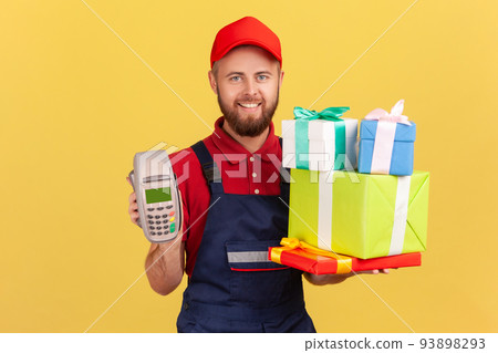 Portrait of positive delighted courier man wearing blue overalls holding stack of present boxes and pos terminal for online paying. Indoor studio shot isolated on yellow background. Portrait of positive delighted courier man wearing blue overalls holding stack of present boxes and pos terminal for online paying. Indoor studio shot isolated on yellow background. 93898293