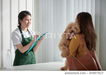 Cheerful female groomer filling form on clipboard when talking to small dog owner Cheerful female groomer filling form on clipboard when talking to small dog owner 93907427