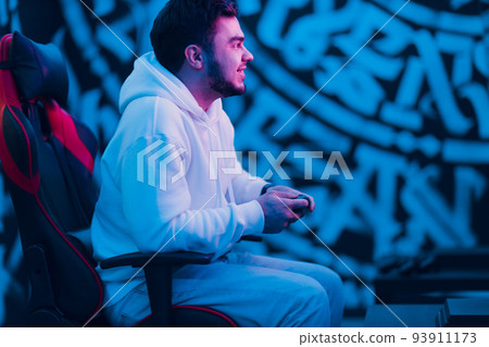 Young man plays on game console with gamepad sitting on chair. Young man plays on game console with gamepad sitting on chair. 93911173