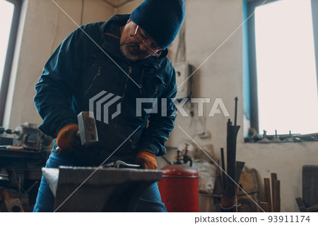 Blacksmith forges and makes metal detail with hammer and anvil at forge. Blacksmith forges and makes metal detail with hammer and anvil at forge. 93911174
