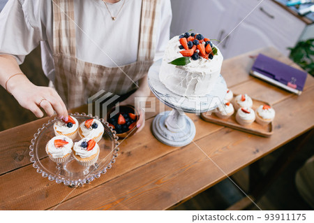 Pastry chef confectioner in beige apron young caucasian woman decorate cake on kitchen table. Cakes cupcakes and sweet dessert Pastry chef confectioner in beige apron young caucasian woman decorate cake on kitchen table. Cakes cupcakes and sweet dessert 93911175