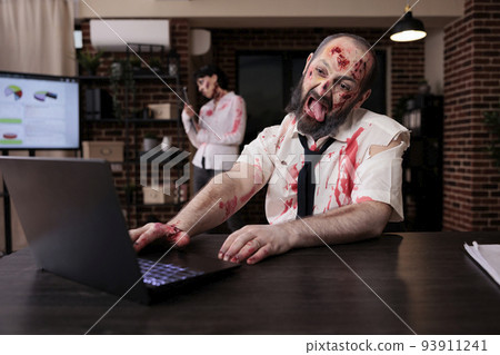 Creepy scary zombie using laptop at desk, looking creepy and horrific working on computer. Terrifying eerie dramatic brain eating monsters sitting in business office, horrible gory walker. 93911241