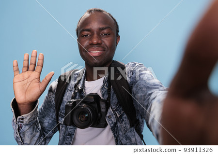 Joyful young adult waving at camera while having professional camera and holiday trip backpack on blue background. Photography enthusiast with DSLR photo device taking picture of himself. 93911326