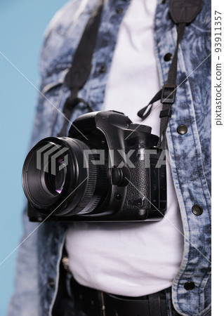 Close up shot of professional photographer having photo device while standing on blue background. Photography enthusiast wearing fashion denim jacket while having modern DSLR camera. 93911357