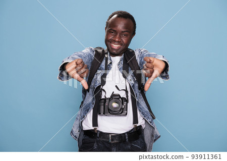 Young african american photography enthusiast showing disapproving hand gesture while standing on blue background. Smiling photographer giving thumbs down negative symbol while having DSLR camera. 93911361