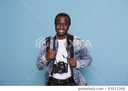 Happy photography enthusiast with DSLR photo device giving okay approve hand symbol. Young photographer showing thumbs up hand gesture while having professional camera on blue background. 93911368