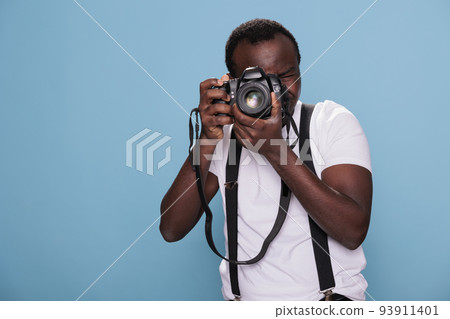 Confident professional photographer having DSLR camera taking picture. African american photography enthusiast taking photo while standing on blue background. Studio shot 93911401
