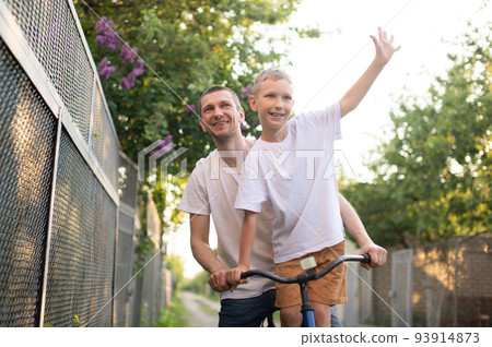 A father rides his son in a white T-shirt on a bicycle. The happy son spread his arms A father rides his son in a white T-shirt on a bicycle. The happy son spread his arms 93914873