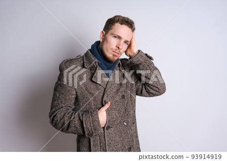 A young bearded man in an autumn coat poses emotionally against a light wall. A young bearded man in an autumn coat poses emotionally against a light wall. 93914919
