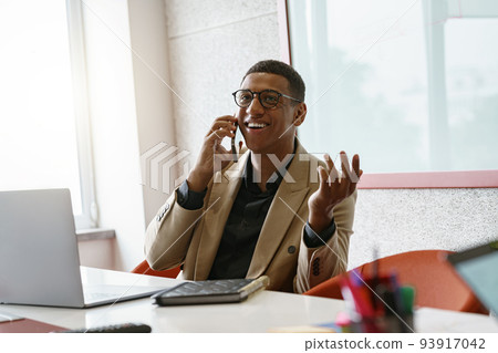 Smiling african businessman in glasses talking phone sitting in cozy coworking Smiling african businessman in glasses talking phone sitting in cozy coworking 93917042