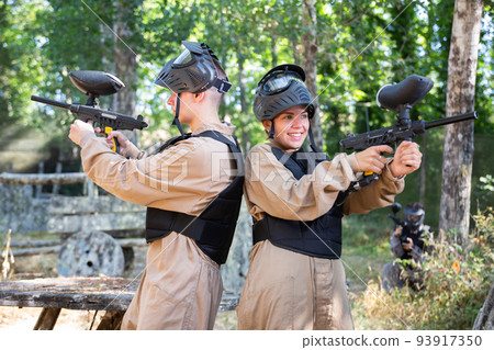 Boy and girl paintball players standing back-to-back and shooting Boy and girl paintball players standing back-to-back and shooting 93917350