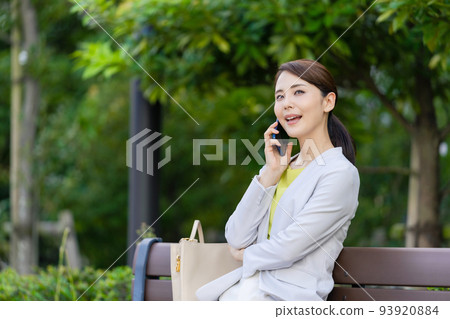 A woman sitting on a park bench and talking on her smartphone A woman sitting on a park bench and talking on her smartphone 93920884