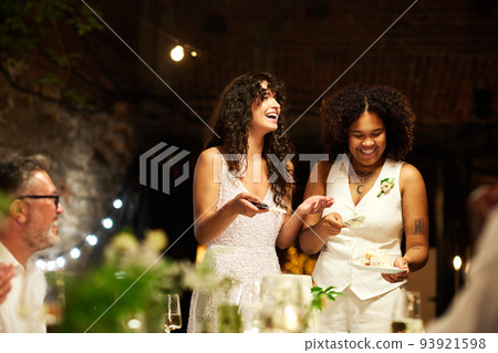 Cheerful young lesbian brides in wedding attire spreading cake among guests 93921598