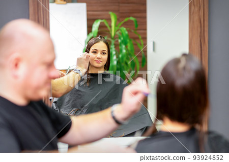 Reflection in the mirror of the young caucasian woman sitting and receiving haircut by male hairdresser at hairdresser salon 93924852