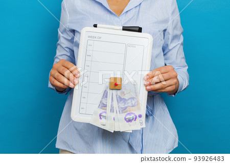 A woman holds a weekly planner for the week and British pounds in her hands in a studio on a blue background. The concept of planning expenses for the week. 93926483