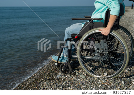 Caucasian woman in a wheelchair on the seashore. Close-up of female hands. 93927537