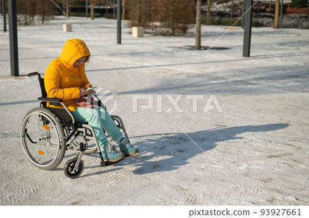 Caucasian woman in a wheelchair uses a smartphone on a walk in the park in winter. Caucasian woman in a wheelchair uses a smartphone on a walk in the park in winter. 93927661