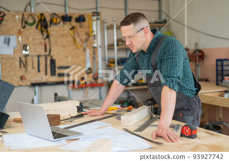 Portrait of a carpenter in protective glasses and work overalls uses a laptop in a workshop. 93927742