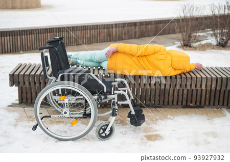 Homeless woman sleeping on a park bench next to a wheelchair. Homeless woman sleeping on a park bench next to a wheelchair. 93927932