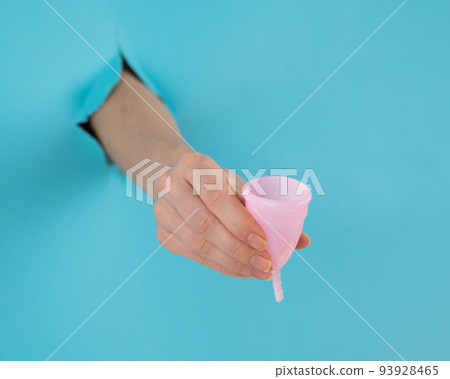 A female hand sticking out of a hole from a blue background holds a pink menstrual cup.  93928465