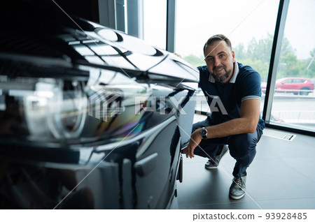 A happy Caucasian man sits near the hood of his new car in a car dealership. A happy Caucasian man sits near the hood of his new car in a car dealership. 93928485