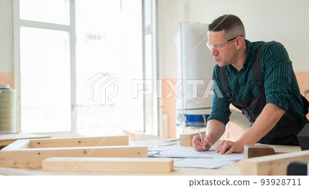 A portrait of a carpenter in goggles and work overalls draws a blueprint for a workshop. 93928711