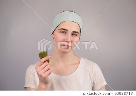 Portrait of a young woman massages her face with a gouache scraper on a white background. 93928717
