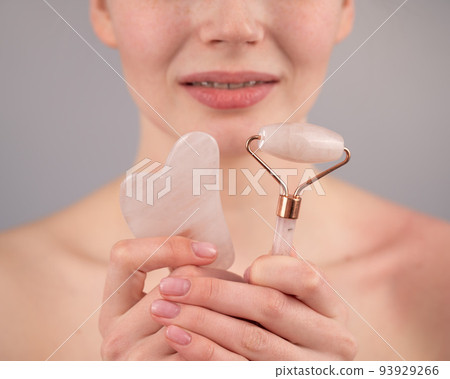 Close-up portrait of a woman with braces on her teeth holding a pink roller massager and a gouache scraper on a white background.  93929266