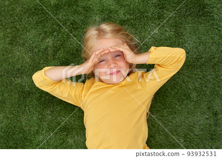Portrait cute blonde girl eyes lying on green grass. Mock up yellow t shirt. Top view. Portrait cute blonde girl eyes lying on green grass. Mock up yellow t shirt. Top view. 93932531