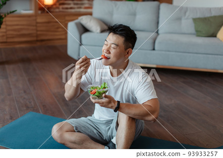 Healthy lifestyle concept. Relaxed asian man eating fresh vegetable salad, sitting on yoga mat after home workout Healthy lifestyle concept. Relaxed asian man eating fresh vegetable salad, sitting on yoga mat after home workout 93933257