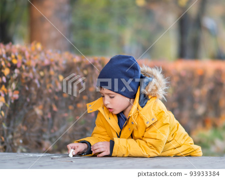 Little boy writes or draws something with chalk on pavement. Creative leisure activity outdoors at fall. Little boy writes or draws something with chalk on pavement. Creative leisure activity outdoors at fall. 93933384