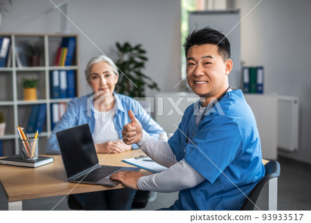 Cheerful adult asian male doctor showing thumb up sign with hand and consultation elderly female patient 93933517