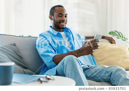Positive black man using smartphone, scrolling news feed in social media app or texting online, sitting on sofa Positive black man using smartphone, scrolling news feed in social media app or texting online, sitting on sofa 93933701