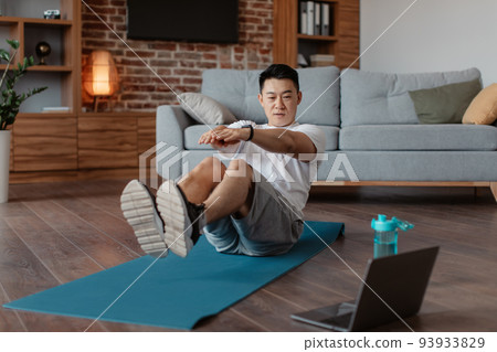 Mature korean man doing exercises for abs on mat on floor in living room interior, watching workout on laptop computer 93933829