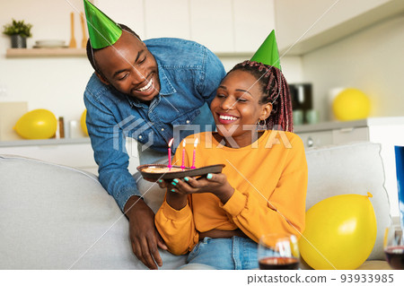 Cheery young black couple in party hats celebrating birthday, woman holding birthday pie with lit candles 93933985