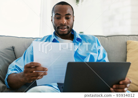 Remote career, freelance job concept. Young african american man using laptop computer and looking through documents Remote career, freelance job concept. Young african american man using laptop computer and looking through documents 93934012