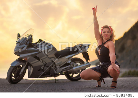A beautiful rocker-woman in leather shorts confidently poses squatting by a motorcycle and showing goat-gesture. The concept of motorbike travel 93934093