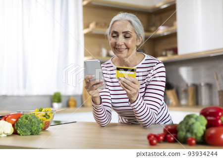 Smiling Senior Woman Using Smartphone And Credit Card While Cooking In Kitchen 93934244