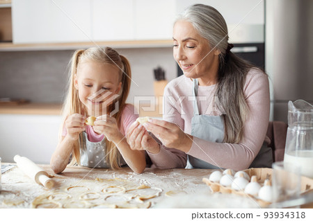 Glad european little girl and old grandmother in aprons make cookies from dough, sculpt dumplings 93934318