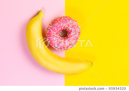 The concept of choice and nutrition. Banana and donut with pink icing on a yellow-pink background. Top view 93934320