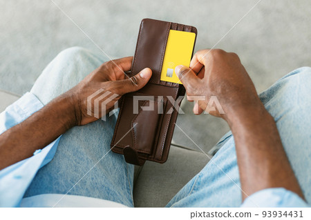 African american man selecting credit card from his card wallet, sitting on couch at home, closeup 93934431