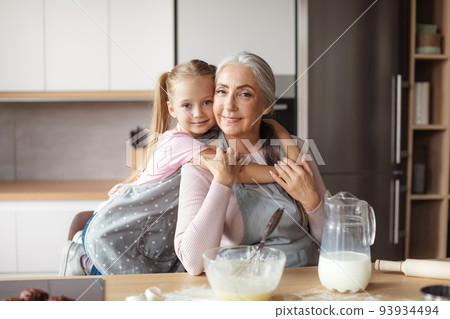 Smiling little girl hugs old woman in apron making dough in kitchen interior 93934494