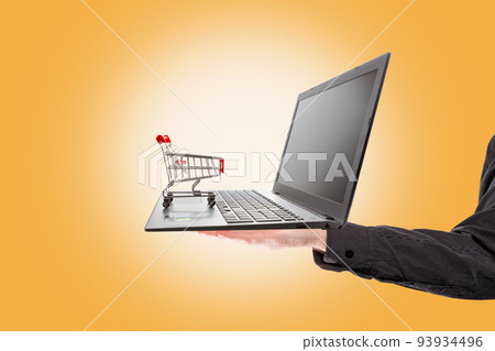 Shopping. A man's hand holds a laptop with a mini grocery cart standing on it. Orange background. The concept of online shopping 93934496