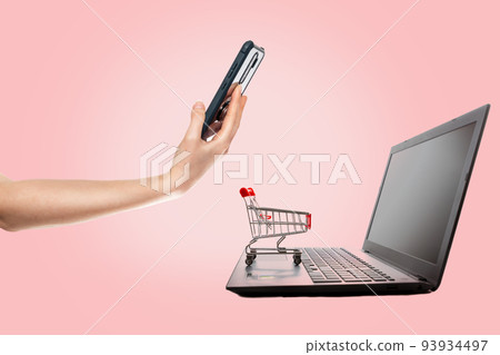 Modern technology. A laptop with a mini grocery cart standing on it, and a woman pays for a purchase with a smartphone. Pink background. The concept of online shopping Modern technology. A laptop with a mini grocery cart standing on it, and a woman pays for a purchase with a smartphone. Pink background. The concept of online shopping 93934497