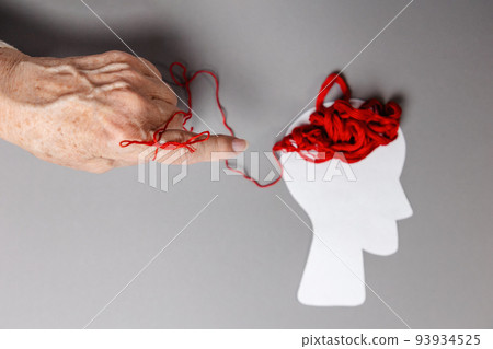 World Alzheimer's Day. A red thread is tied on the index finger of the senior woman's hand. Tangled threads symbolizing the brain on the defocused paper silhouette of the head 93934525