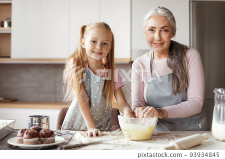 Granddaughter and grandma prepare cookies. Glad european little girl and elderly female in aprons make dough 93934845