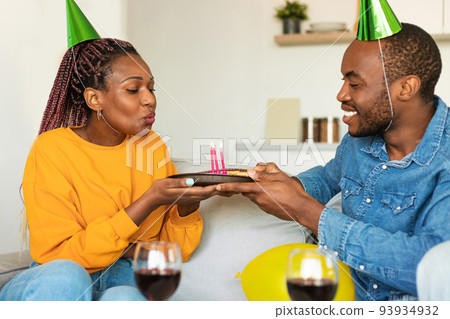 Cheery black couple in party hats celebrating birthday, woman blowing candles on pie, drinking wine at home 93934932