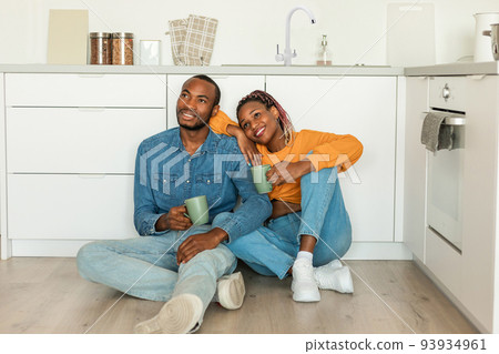 Romantic black couple drinking coffee and looking away, sitting on floor in kitchen, enjoying morning at home Romantic black couple drinking coffee and looking away, sitting on floor in kitchen, enjoying morning at home 93934961