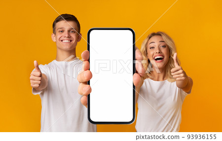 Cheerful excited young caucasian couple with open mouth show smartphone with blank screen and thumbs up Cheerful excited young caucasian couple with open mouth show smartphone with blank screen and thumbs up 93936155