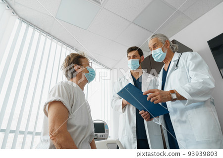 Doctors in masks explain to a sick patient in a wheelchair her diagnosis and treatment 93939803